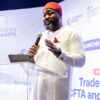 Man in white traditional outfit and red hat speaks into a microphone at a clear podium during a conference with banners in the background.