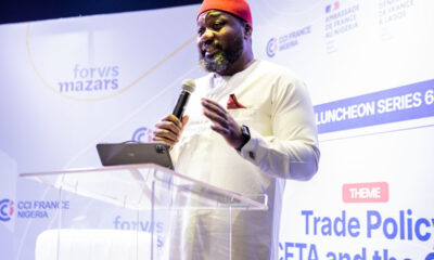 Man in white traditional outfit and red hat speaks into a microphone at a clear podium during a conference with banners in the background.