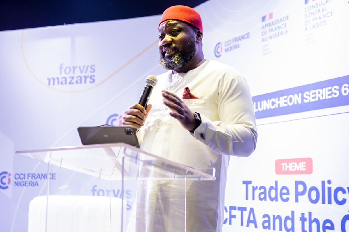 Man in white traditional outfit and red hat speaks into a microphone at a clear podium during a conference with banners in the background.
