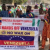 Group of protesters in Nigeria holding a banner that reads 'Venezuela Solidarity Campaign in Nigeria' and 'US HANDS OFF VENEZUELA' with Venezuelan flags nearby, smiling faces visible on a sunny street.