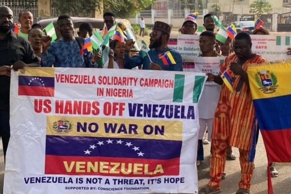 Group of protesters in Nigeria holding a banner that reads 'Venezuela Solidarity Campaign in Nigeria' and 'US HANDS OFF VENEZUELA' with Venezuelan flags nearby, smiling faces visible on a sunny street.