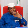 Man in a white cap and blue traditional outfit sits in a red chair, gesturing with his right hand.
