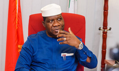Man in a white cap and blue traditional outfit sits in a red chair, gesturing with his right hand.