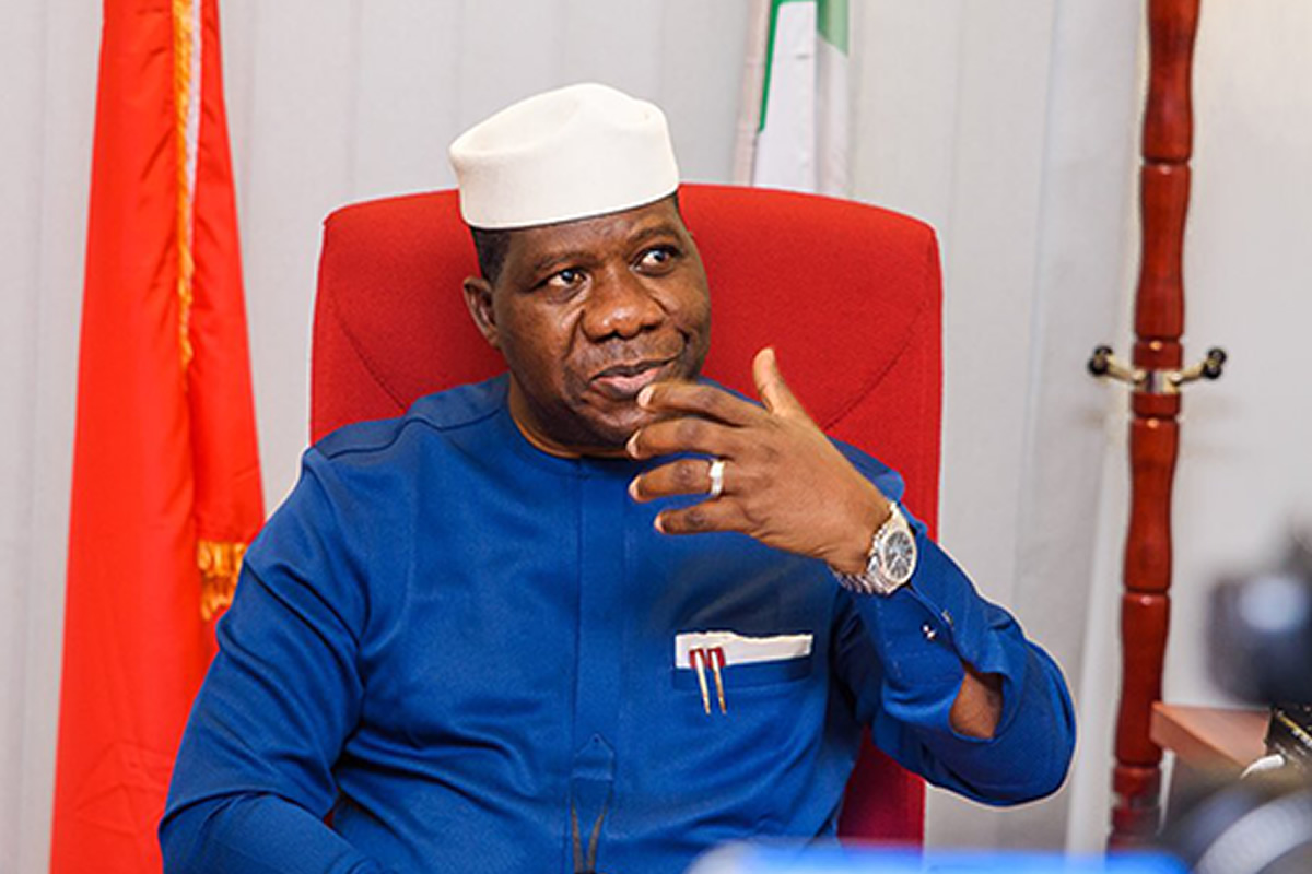 Man in a white cap and blue traditional outfit sits in a red chair, gesturing with his right hand.