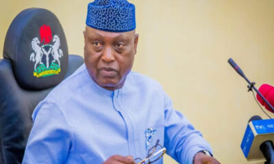 Man in a light blue traditional outfit and patterned blue cap sits at a desk during a press briefing, holding his glasses with TVC News microphones nearby.