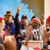 Group of people at a political rally raising victory signs and speaking into microphones on a stage with a crowd in front.
