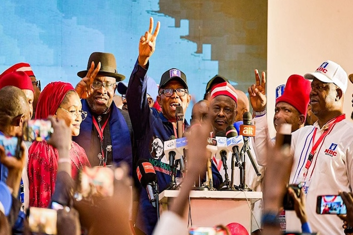 Group of people at a political rally raising victory signs and speaking into microphones on a stage with a crowd in front.