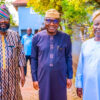 Three smiling men in traditional West African attire stand outdoors in a sunny courtyard, posing for a photo.