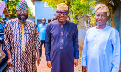 Three smiling men in traditional West African attire stand outdoors in a sunny courtyard, posing for a photo.