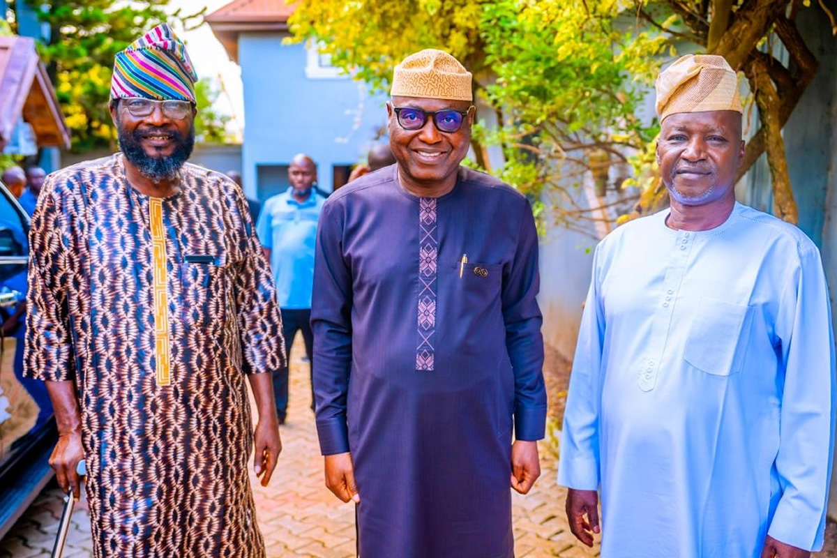 Three smiling men in traditional West African attire stand outdoors in a sunny courtyard, posing for a photo.