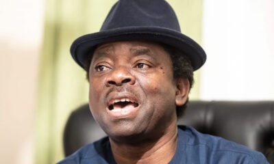 Man wearing a dark fedora hat and blue shirt, mouth open as if singing or speaking.