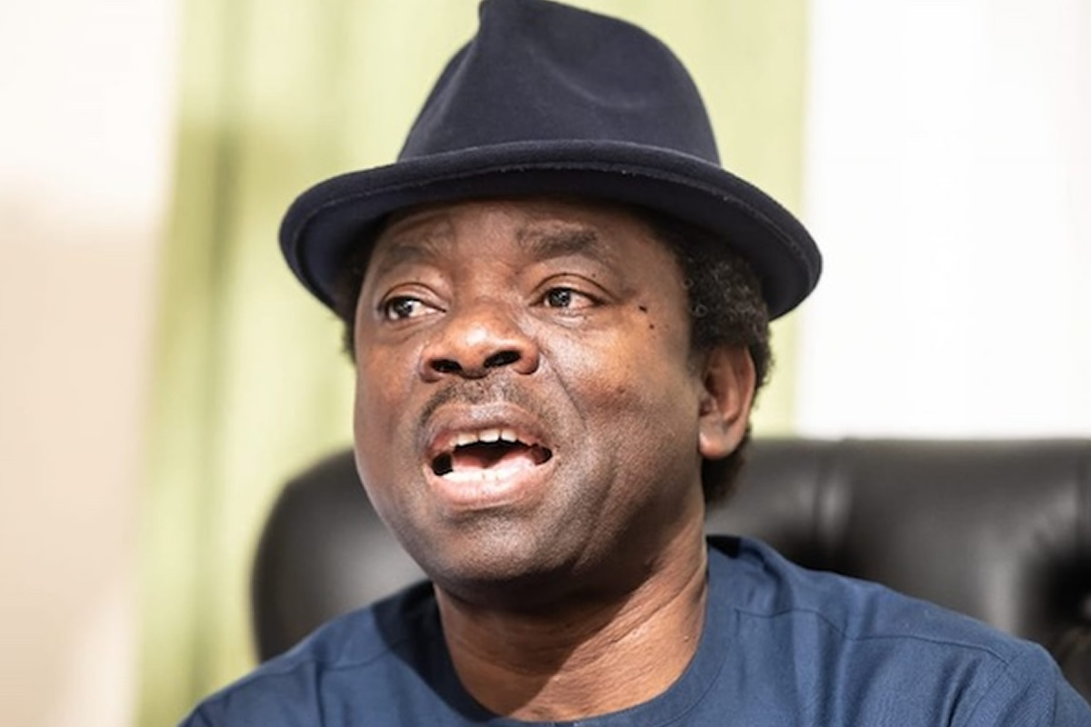 Man wearing a dark fedora hat and blue shirt, mouth open as if singing or speaking.