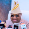 Man in beige traditional attire and striped hat speaks at a press conference with multiple microphones in front of him, wearing sunglasses.