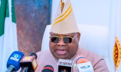 Man in beige traditional attire and striped hat speaks at a press conference with multiple microphones in front of him, wearing sunglasses.