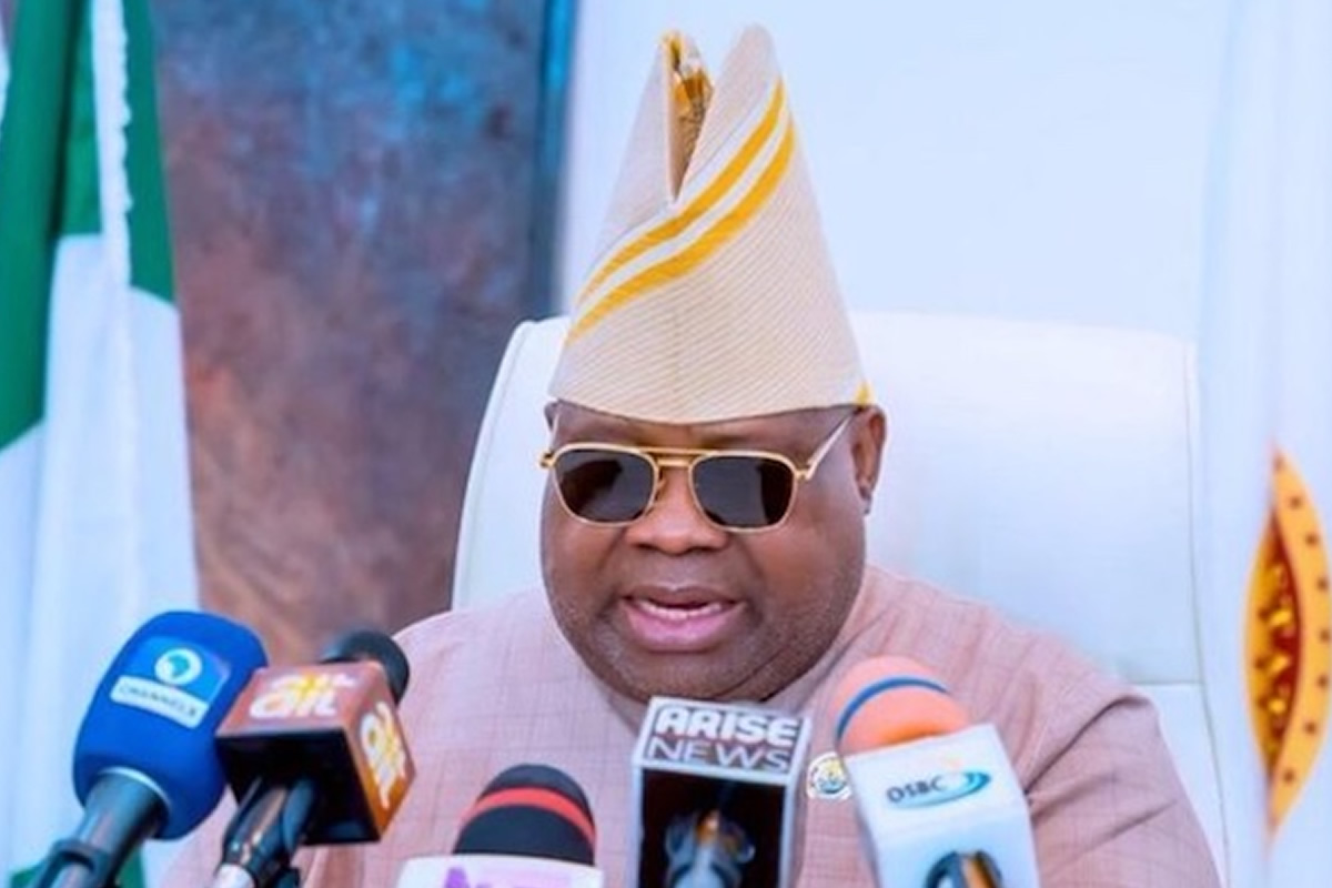 Man in beige traditional attire and striped hat speaks at a press conference with multiple microphones in front of him, wearing sunglasses.