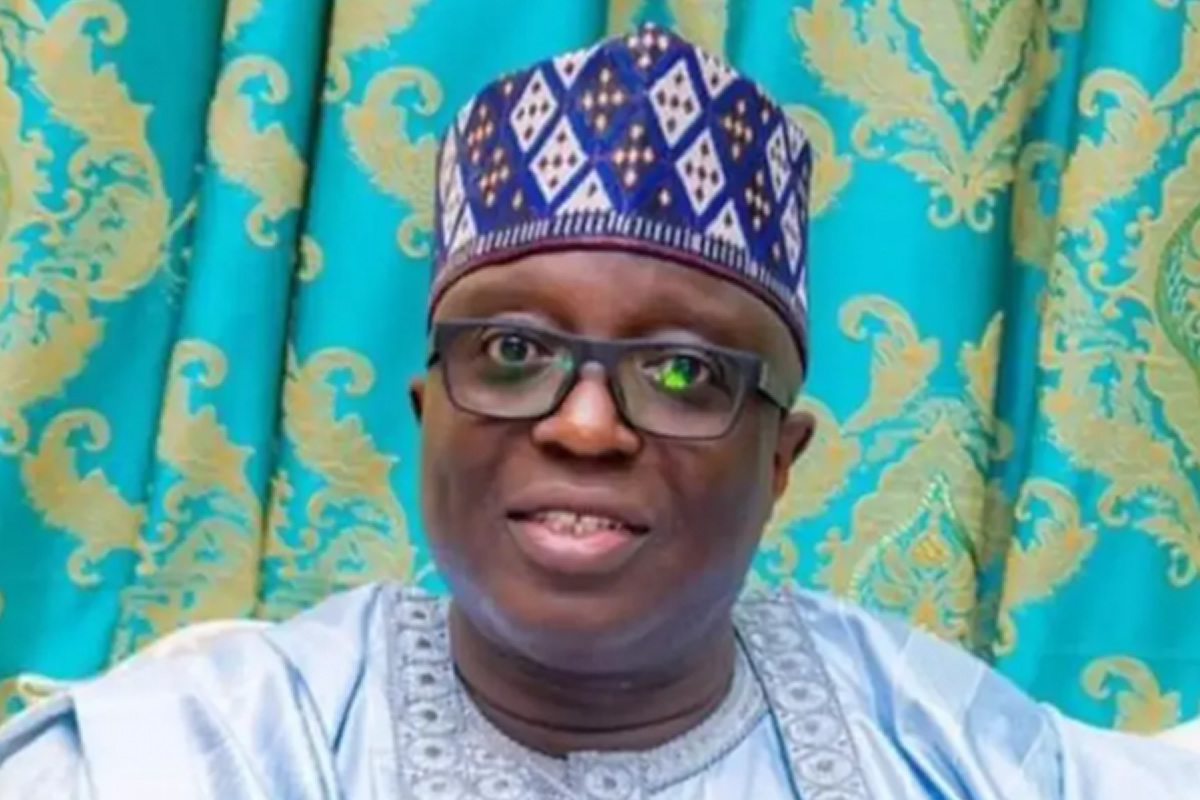 Smiling man wearing a blue patterned cap and glasses, posed in front of turquoise and gold curtains.