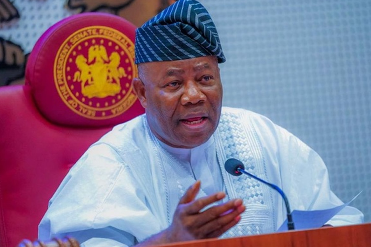 Man in traditional white attire speaking into a microphone at a podium, with a red ceremonial chair featuring a gold emblem behind him.