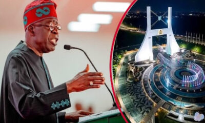 A man in traditional attire speaks at a podium, with a circular inset on the right showing a nighttime view of a futuristic white arch monument and illuminated surrounding plaza.