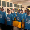 Group of children in blue UNICEF World Children's Day shirts stand in a line at an indoor event hall, some wearing hijabs, ready for a ceremony.