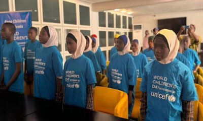 Group of children in blue UNICEF World Children's Day shirts stand in a line at an indoor event hall, some wearing hijabs, ready for a ceremony.
