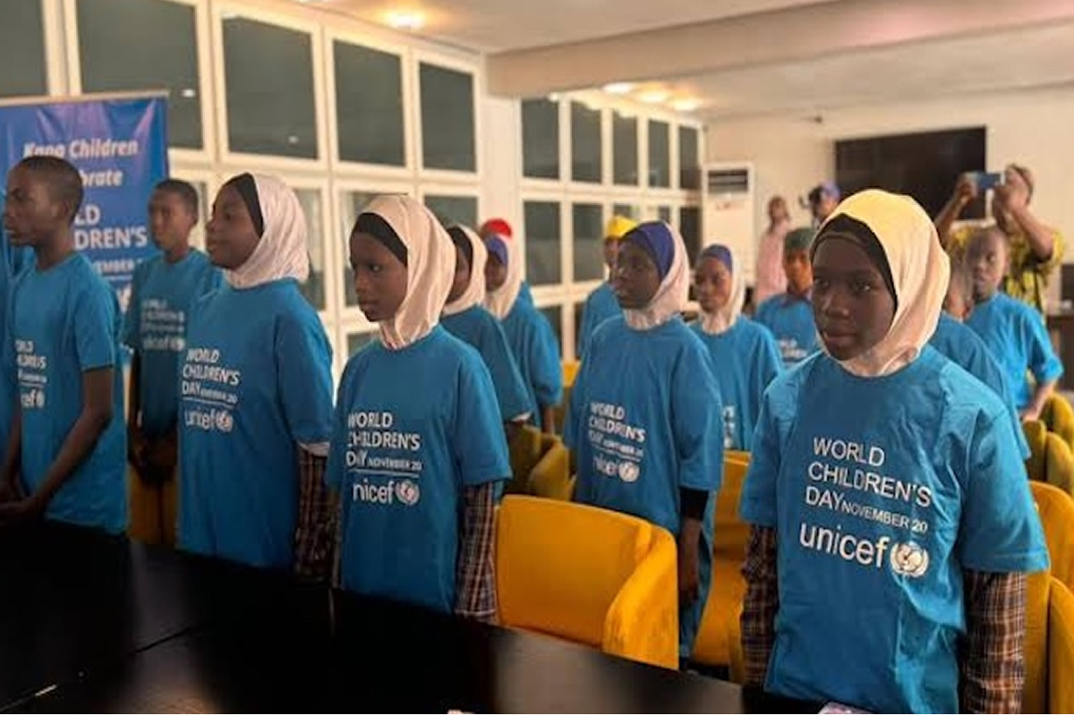 Group of children in blue UNICEF World Children's Day shirts stand in a line at an indoor event hall, some wearing hijabs, ready for a ceremony.