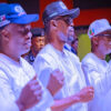 Group of men in white traditional outfits standing in a line, fists raised in a ceremonial gesture during an event.