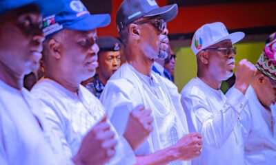 Group of men in white traditional outfits standing in a line, fists raised in a ceremonial gesture during an event.