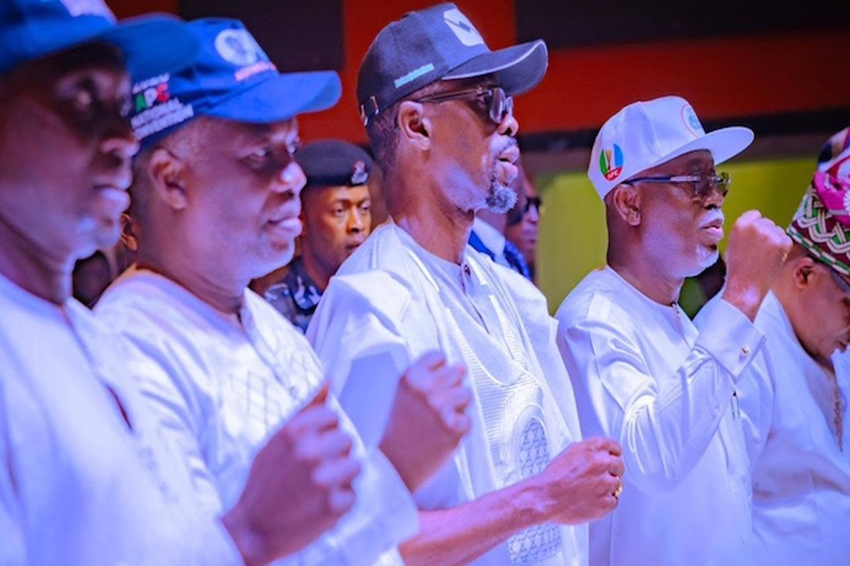 Group of men in white traditional outfits standing in a line, fists raised in a ceremonial gesture during an event.