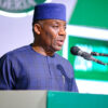Man in blue traditional attire and hat speaking at a podium with a microphone during a formal event.