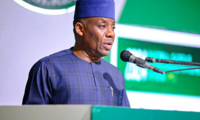 Man in blue traditional attire and hat speaking at a podium with a microphone during a formal event.