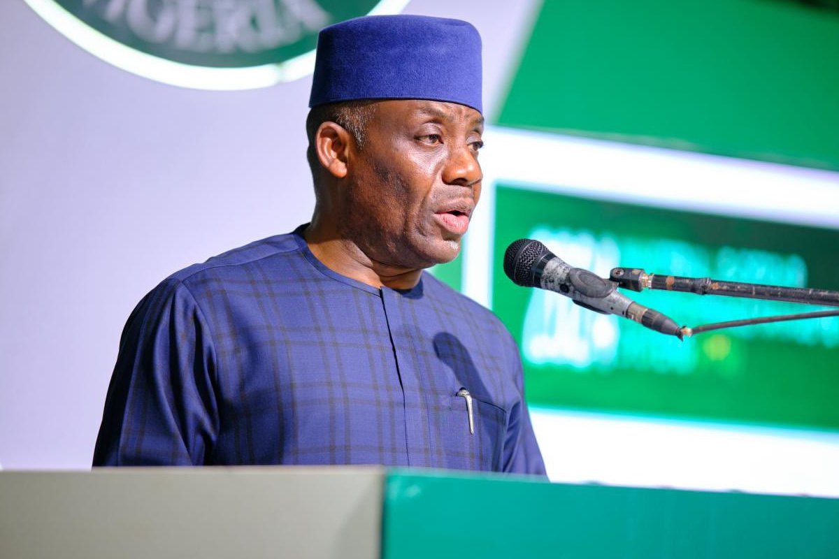 Man in blue traditional attire and hat speaking at a podium with a microphone during a formal event.