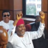 Smiling man in white traditional attire and red cap raises both arms in a meeting room while others sit nearby.