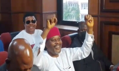 Smiling man in white traditional attire and red cap raises both arms in a meeting room while others sit nearby.