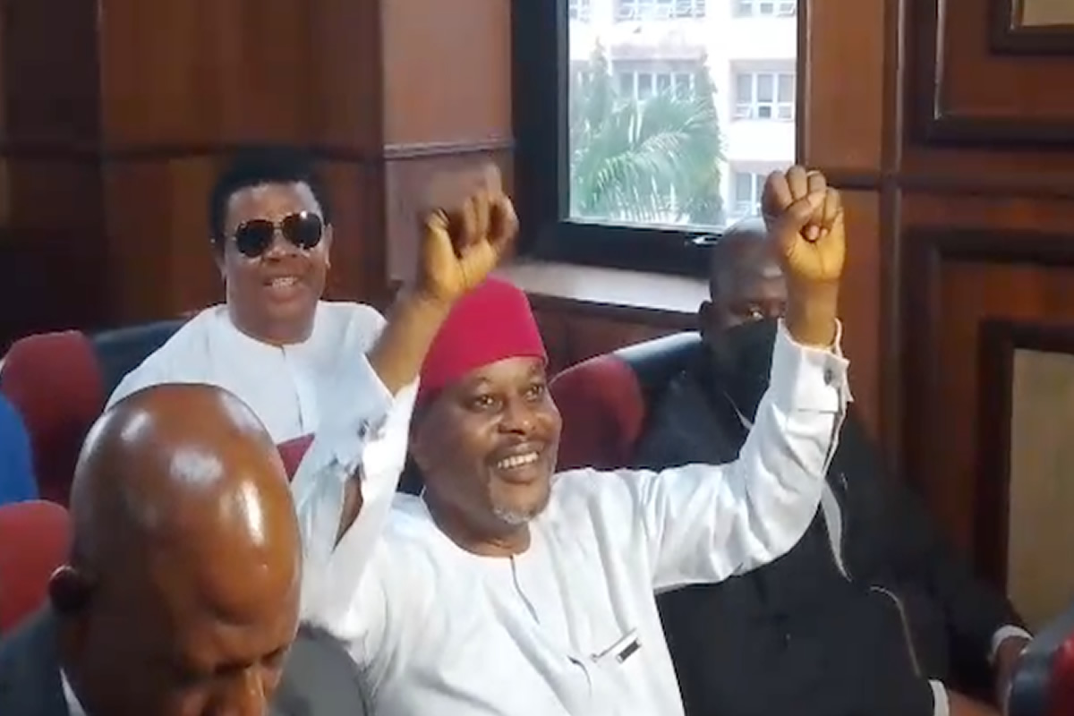 Smiling man in white traditional attire and red cap raises both arms in a meeting room while others sit nearby.