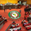 Wide shot of the ECOWAS Parliament chamber with red seats and a large circular ECOWAS emblem on the carpet, surrounded by officials and desks.",