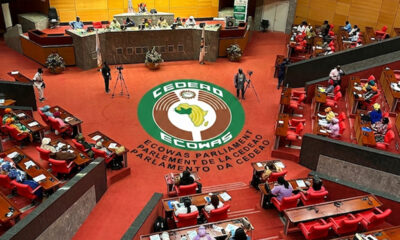 Wide shot of the ECOWAS Parliament chamber with red seats and a large circular ECOWAS emblem on the carpet, surrounded by officials and desks.",