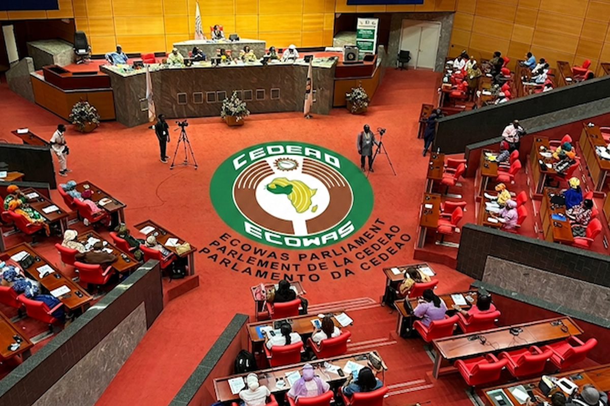 Wide shot of the ECOWAS Parliament chamber with red seats and a large circular ECOWAS emblem on the carpet, surrounded by officials and desks.",