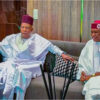 Two men in white Yoruba agbada sit on a sofa, holding hands and facing forward in a living room setting.
