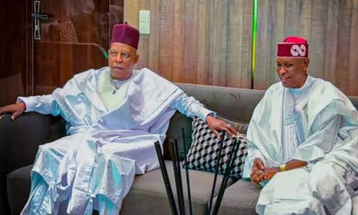 Two men in white Yoruba agbada sit on a sofa, holding hands and facing forward in a living room setting.