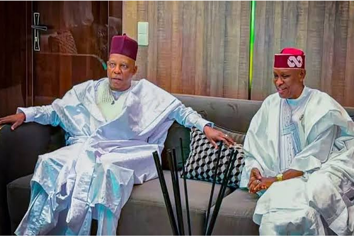 Two men in white Yoruba agbada sit on a sofa, holding hands and facing forward in a living room setting.