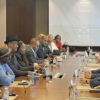Diverse group of professionals seated around a long conference table in a meeting room, facing a projector screen that reads 'Welcome'. They have laptops, papers, and water bottles on the table.