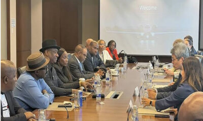 Diverse group of professionals seated around a long conference table in a meeting room, facing a projector screen that reads 'Welcome'. They have laptops, papers, and water bottles on the table.