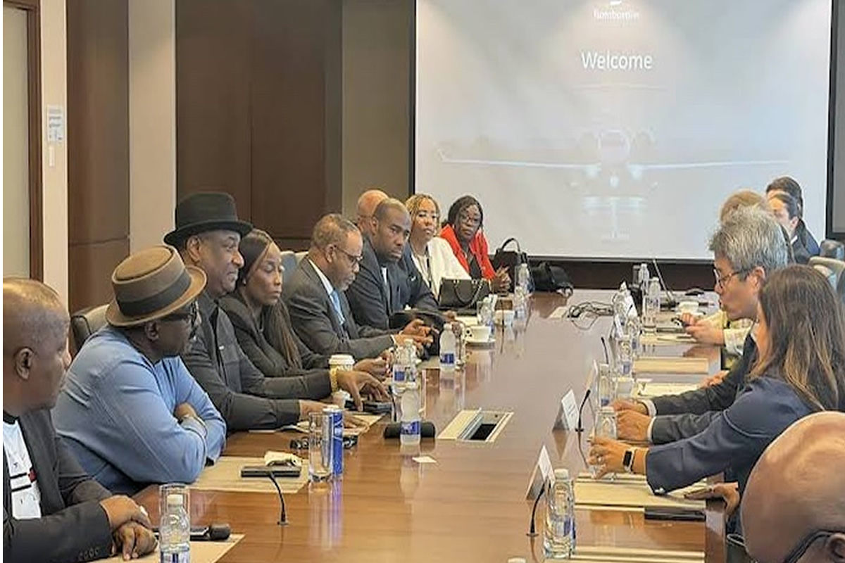 Diverse group of professionals seated around a long conference table in a meeting room, facing a projector screen that reads 'Welcome'. They have laptops, papers, and water bottles on the table.