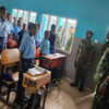 Classroom scene with students in blue uniforms standing beside desks as armed personnel observe at the front.