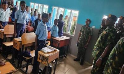 Classroom scene with students in blue uniforms standing beside desks as armed personnel observe at the front.