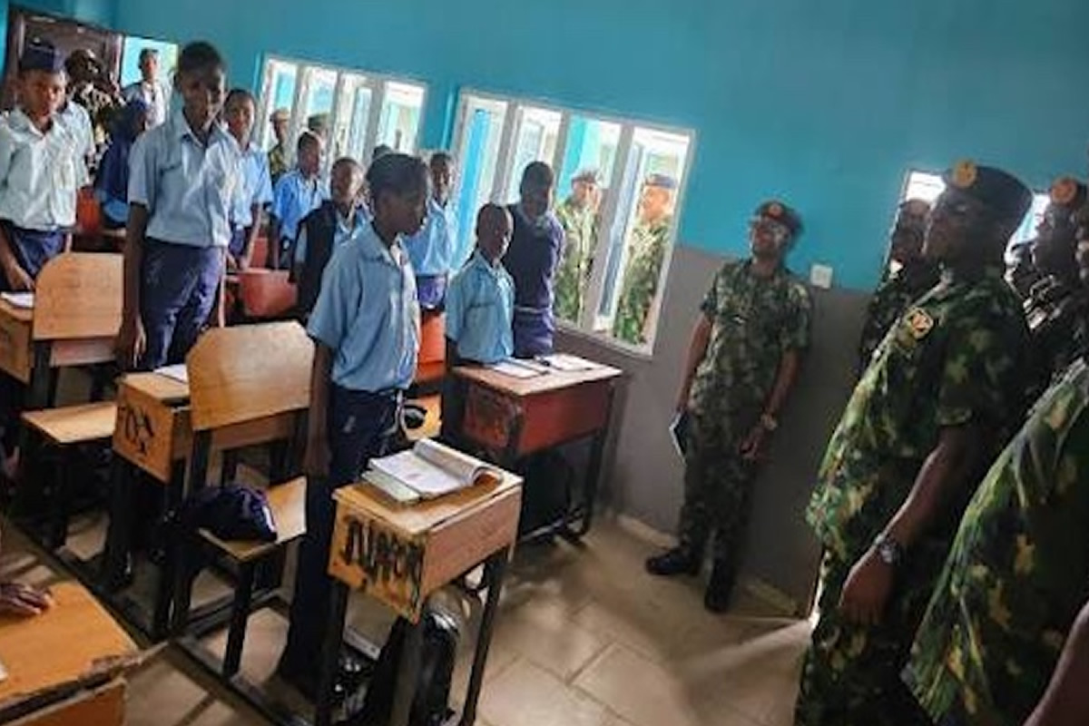 Classroom scene with students in blue uniforms standing beside desks as armed personnel observe at the front.