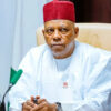 Older man in a white traditional outfit and red cap sits at a desk with a green microphone, looking serious in a formal meeting setting.