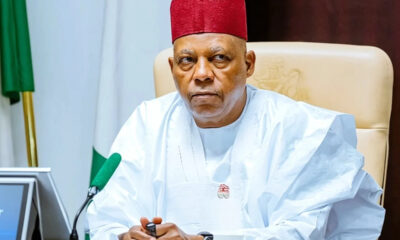 Older man in a white traditional outfit and red cap sits at a desk with a green microphone, looking serious in a formal meeting setting.