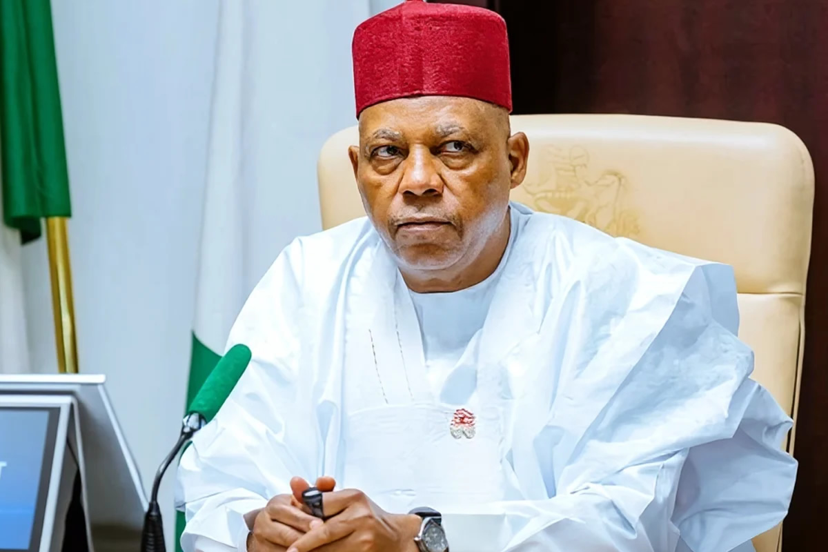 Older man in a white traditional outfit and red cap sits at a desk with a green microphone, looking serious in a formal meeting setting.