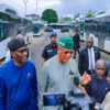 Group of Nigerian officials, including a man in a green cap, addressing reporters beside waiting buses at a transport yard.